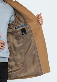 Beige Mantel mit einer glatten, strukturierten Außenseite. Innen befindet sich ein glänzendes braunes Futter, eine blau gemusterte Tasche und ein Markenetikett.