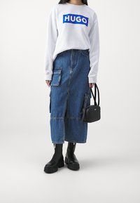 Felpa bianca con testo "HUGO" blu, abbinata a una gonna midi in denim con tasche laterali e uno spacco frontale. Stivali neri alla caviglia e una piccola borsa nera.
