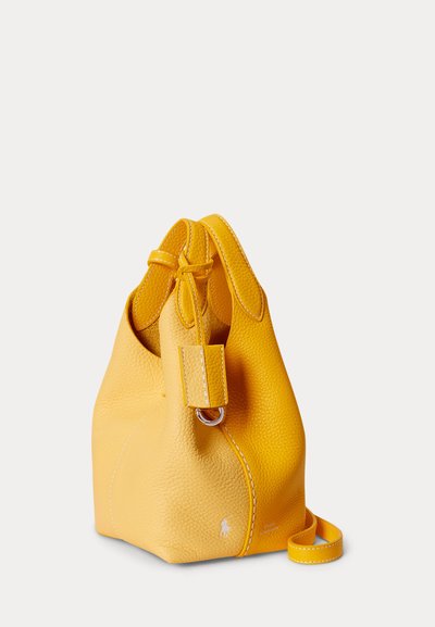 Gul lædertaske i spandform med top-håndtag, synlige syninger og en lille sølvlogo nær bunden på en ensfarvet baggrund.