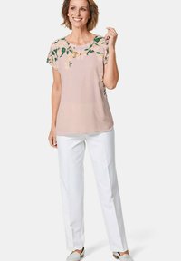Helles rosa Blusenshirt mit floralem Stickerei an den Schultern, kombiniert mit weißen Hosen und hellen Slippern. Glatte Stoffqualität, entspannte Passform.