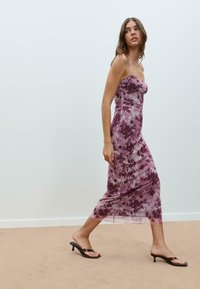 Robe maxi tie-dye violet avec un corsage ajusté, des bretelles fines et une silhouette fluide, associée à des sandales à talons noires.