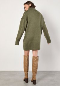 Jersey de punto oversize en verde oliva con cuello alto y puños acanalados, combinado con botas de cuero de color tostado hasta la rodilla con tacones anchos.