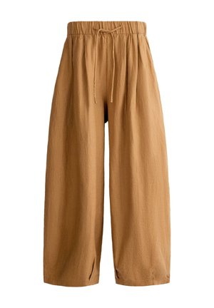 Žena u uskoj bijeloj majici kratkih rukava, rastopljenim smeđim pantalonama s trakicom za vezanje i crnim sandalama, stoji na svijetlom pločastom podu.