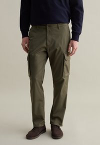 Olivgrüne Cargo-Hosen mit mehreren Seitentaschen, einer lockeren Passform und einem Reißverschluss. Kombiniert mit dunkelbraunen Wildlederschuhen und einem marineblauen Pullover.