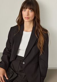 Černé na míru šité sako s jedním knoflíkem, nošené přes bílý žebrovaný top. Dlouhé vlnité vlasy, drží černé psaníčko. Jednoduchý, moderní design.