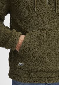 Olive green fleece pullover with textured surface, half-zip front, adjustable drawstring, and kangaroo pocket; brand label visible on hem.