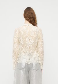 Blouse en dentelle à manches longues de couleur blanc cassé, présentant un ourlet festonné et une fermeture boutonnée au dos, associée à un pantalon large argenté.