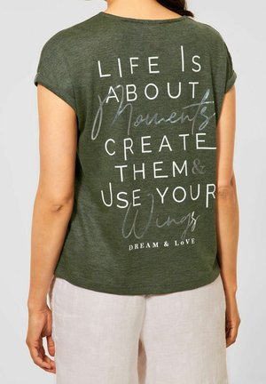 Vrouw draagt een olijfgroene t-shirt met witte tekst op de achterkant: "Het leven draait om momenten, creëer ze & gebruik je vleugels, droom & houd van."