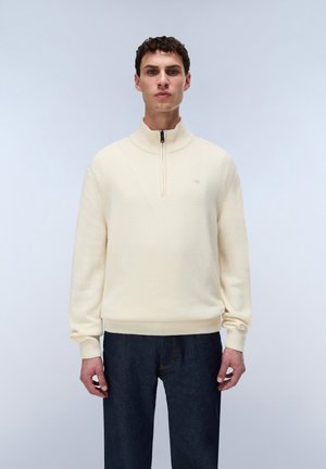 Mand iført en cremefarvet halv-zip sweater og mørkeblå jeans står mod en ensfarvet lysegrå baggrund.