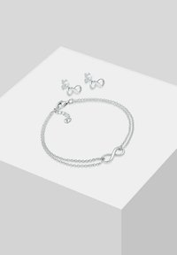 Bracciale infinito in argento con due catene e chiusura a moschettone; gli orecchini a bottone coordinati presentano anch'essi un design infinito. Finitura in metallo liscia e lucida.