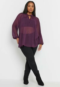 Blusa viola trasparente con maniche lunghe e scollo a cravatta. Presenta un motivo a pois. Abbinata a jeans skinny neri e stivali neri alla caviglia.