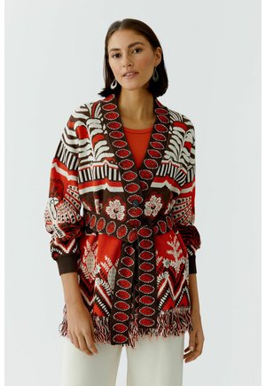 Frau trägt eine gemusterte rote, braune und weiße Strickjacke mit Gürtel, floralem und geometrischem Design, kombiniert mit weißen Hosen und teardrop-förmigen Ohrringen.