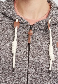 Felpa a maglia marrone e grigia con cappuccio, caratterizzata da un cordino in corda beige, inserti in pelle e una cerniera nera. Esibita con un colletto rosa chiaro sotto.