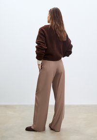 Brauner Strickpullover mit herabgesetzten Schultern, kombiniert mit beigen maßgeschneiderten Hosen. Das Outfit zeichnet sich durch eine lockere Passform aus und betont den Komfort.