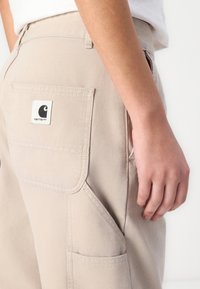 Beige lerretsbukser har en stor lomme på baksiden, synlig søm og en firkantet merkeetikett med logo. Glatt tekstur og avslappet passform.