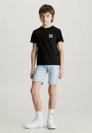 Camiseta de algodón negra con logo blanco, pantalones cortos de mezclilla azul claro, zapatillas blancas y calcetines cortos blancos. Ajuste casual con mangas cortas y pantalones cortos hasta la rodilla.