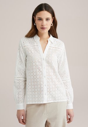 Blusa bianca con bottoni, caratterizzata da ricami ad occhiello, scollatura a V e maniche lunghe con polsini arricciati. Tessuto leggero e vestibilità comoda.