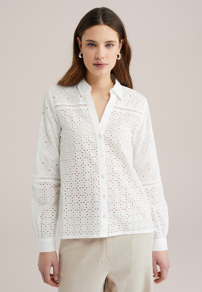Blusa bianca con bottoni, caratterizzata da ricami ad occhiello, scollatura a V e maniche lunghe con polsini arricciati. Tessuto leggero e vestibilità comoda.