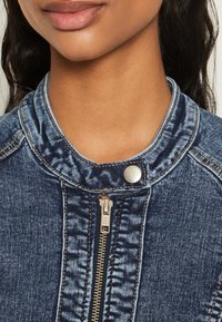 Jeansjacka med ståkrage, som har en silver tryckknapp och en guldfärgad dragkedja. Tyget har en strukturerad blå yta.