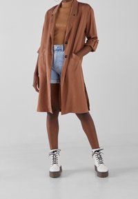 Brązowy, oversize trencz z dwoma przednimi kieszeniami, noszony na beżowej koszuli i dżinsowych szortach; w połączeniu z masywnymi, białymi botkami na sznurówki.