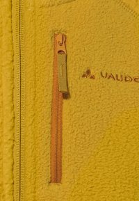 Giacca in pile gialla con superficie testurizzata, tasca con zip arancione e logo a contrasto ricamato sul tessuto in colori abbinati.