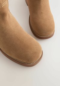 Tādas ķirzakas krāsas zamšādas potīšu zābaki ar apaļu purngalu, kontrastējošu šuvēm apšūtu zoli un gludu virsmu.