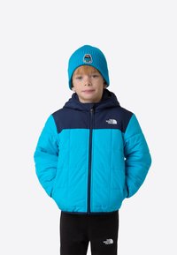 Pikowana kurtka w jasnoniebieskim kolorze z ciemnoniebieskimi akcentami, wyposażona w zamek błyskawiczny z przodu, kaptur i widoczne logo marki. Noszona z dopasowaną czapką typu beanie.