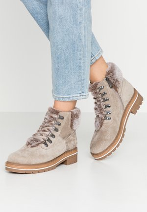 Lysebrune ankelstøvler i ruskind med pelskant, metaløjer, sidelommer med lynlås, båret med lyseblå opkrogede jeans på et hvidt gulv.