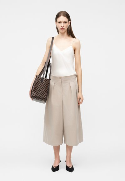 Γυναίκα στέκεται φορώντας μπεζ παντελόνι με φαρδιά, κομμένα πόδια, λευκό αμάνικο τοπ, μαύρα μυτερά παπούτσια και κρατά τσαντάκι ώμου καφέ με σχέδιο.
