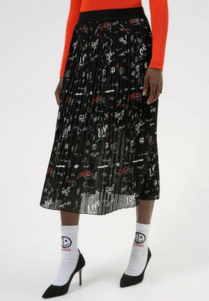 Czarna plisowana spódnica midi z białym i czerwonym odręcznym tekstem oraz symbolami, noszona z białymi skarpetkami z logo i czarnymi butami na wysokim obcasie.