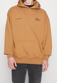 Sweat à capuche marron surdimensionné en matériau doux, avec une poche avant et des poignets côtelés. Texte imprimé sur la poitrine et la manche en couleurs contrastantes.