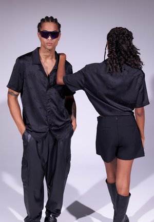 Camicia nera in satin a maniche corte con motivo floreale, abbinata a pantaloni corti neri attillati e stivali al ginocchio. Modella con occhiali da sole.