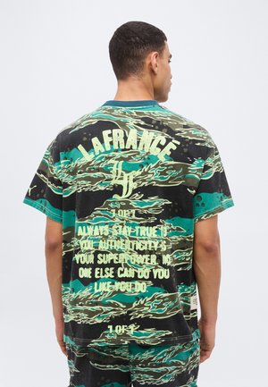 Rövid hajú férfi hátulról, zöld-fekete mintás ingben és rövidnadrágban, az ing hátán nagy fehér felirattal.