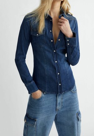Femme portant une chemise en denim bleu foncé boutonnée et un jean cargo en denim bleu clair, une main dans la poche et l'autre ajustant le col.