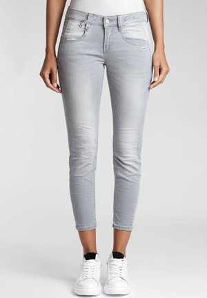 Vrouw draagt lichtgrijze skinny jeans, korter boven de enkels, en witte sneakers, staand tegen een effen achtergrond.