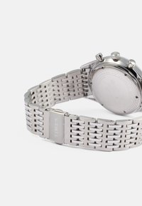 Srebrna kronograf ura iz nerjavečega jekla z mrežasto naramnico. Ima eleganten dizajn, okroglo ohišje in teksturirano sponko z vgraviranim napisom "TIMEX".