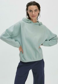 Hellgrüner Fleece-Hoodie mit entspannter Passform, Kordelzugkapuze, gerippten Bündchen und glatter Textur, kombiniert mit dunkelblauen Hosen.
