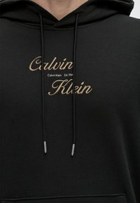 Μαύρο φούτερ με μπεζ κεντημένο κείμενο "Calvin Klein" και μαύρα κορδόνια με μεταλλικά άκρα, φορεμένο από άτομο.