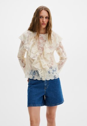 Frau mit langem Haar trägt eine transparente cremefarbene Bluse mit Blumenmuster und Rüschen sowie blaue Jeansshorts vor einem schlichten weißen Hintergrund.