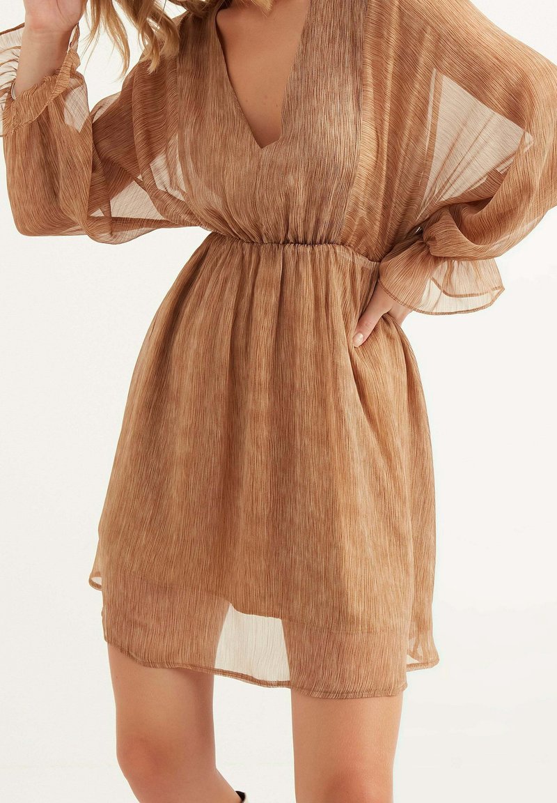 Femme portant une robe marron transparente jusqu'aux genoux avec de longues manches amples et une taille cintrée, posant avec une main sur la hanche.