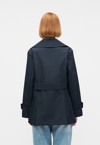 Άτομο με κοντά κόκκινα μαλλιά, που φοράει σκούρο μπλε σακάκι και ανοιχτό μπλε τζιν, εμφανίζεται από πίσω σε απλό φόντο.