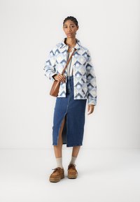 Chaqueta texturizada azul y blanca con patrón floral, combinada con una falda de mezclilla con una abertura frontal, zapatos de plataforma marrones y un bolso marrón.