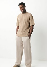 T-shirt en coton beige à manches courtes et coupe décontractée, associée à un pantalon beige clair. Le mannequin se tient les mains dans les poches.