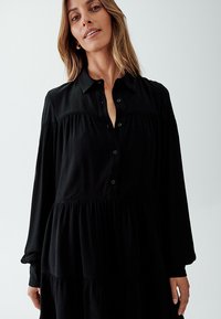 Abito nero a strati con chiusura a bottoni, maniche lunghe, tessuto testurizzato e colletto. Design semplice con dettagli di pieghe sottili.