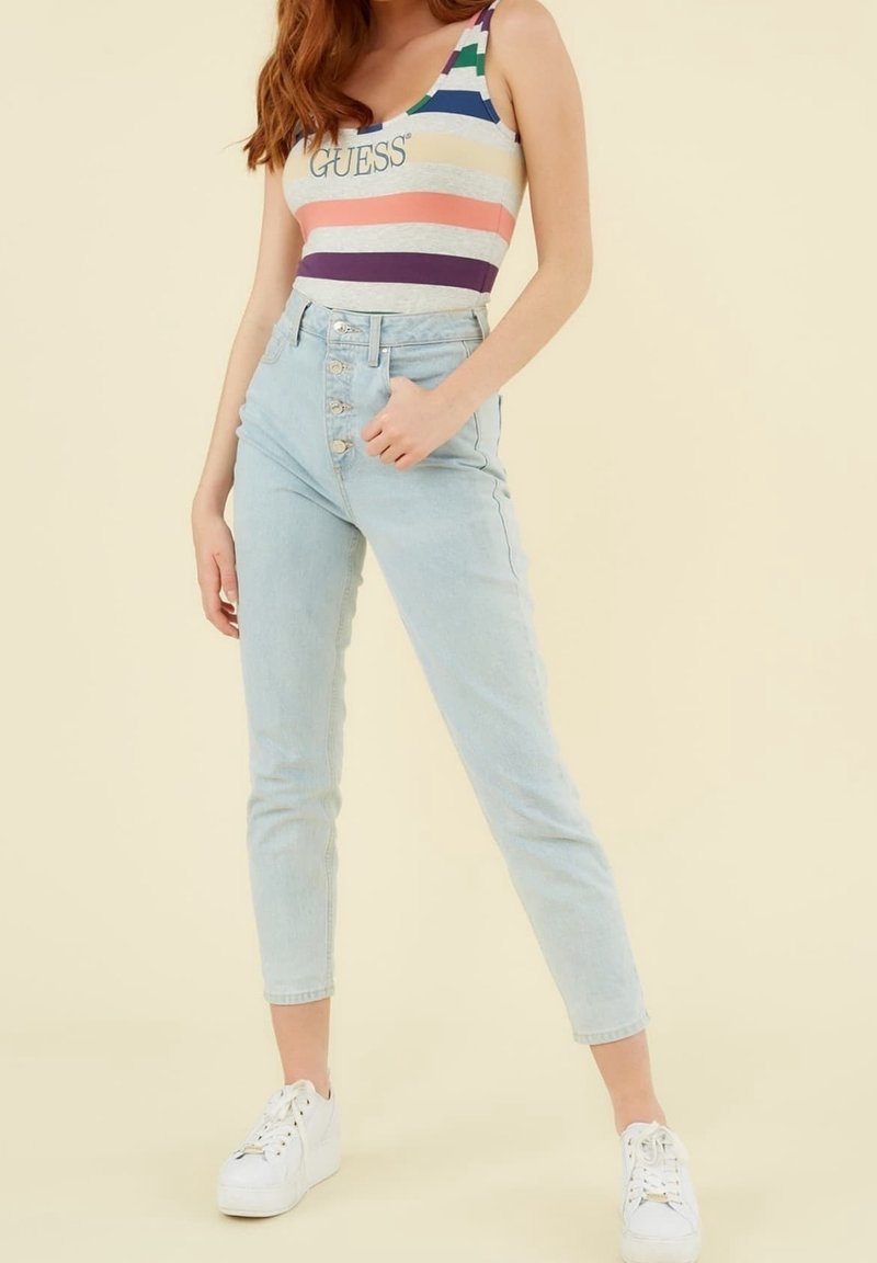 Femme portant un débardeur rayé multicolore avec le texte "GUESS", un jean taille haute bleu clair avec boutons, et des baskets blanches à plateforme.