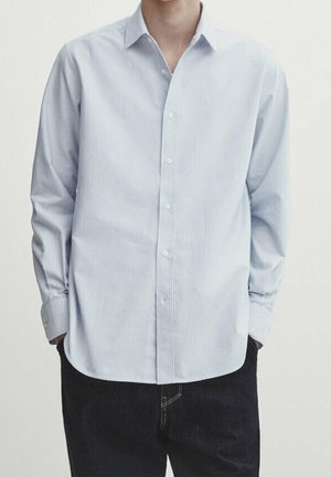 Homme portant une chemise bleu clair à fines rayures, boutonnée à manches longues, non rentrée sur un pantalon noir, les mains dans les poches.