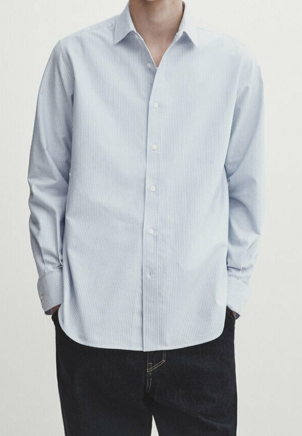 Homme portant une chemise bleu clair à fines rayures, boutonnée à manches longues, non rentrée sur un pantalon noir, les mains dans les poches.