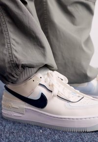 Fehér bőr Nike Air Force 1 sportcipő, tengerészkék swoosh-zal, textúrázott részletekkel és áttetsző talppal. Szürke nadrág részben látható.