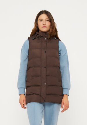 Kvinde iført en brun dunvest over en lyseblå hættetrøje og matchende sweatpants, stående med fronten fremad mod en ensfarvet baggrund.