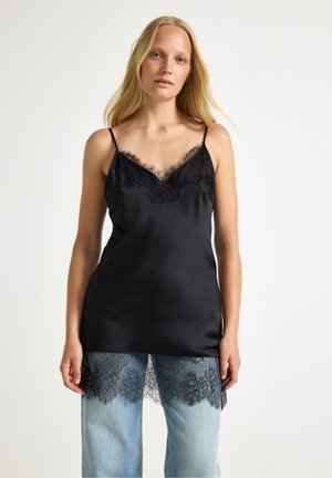 Donna che indossa una camisole di raso nero con bordi in pizzo sopra jeans azzurri chiari con dettagli in pizzo, in piedi davanti a uno sfondo bianco uniforme.